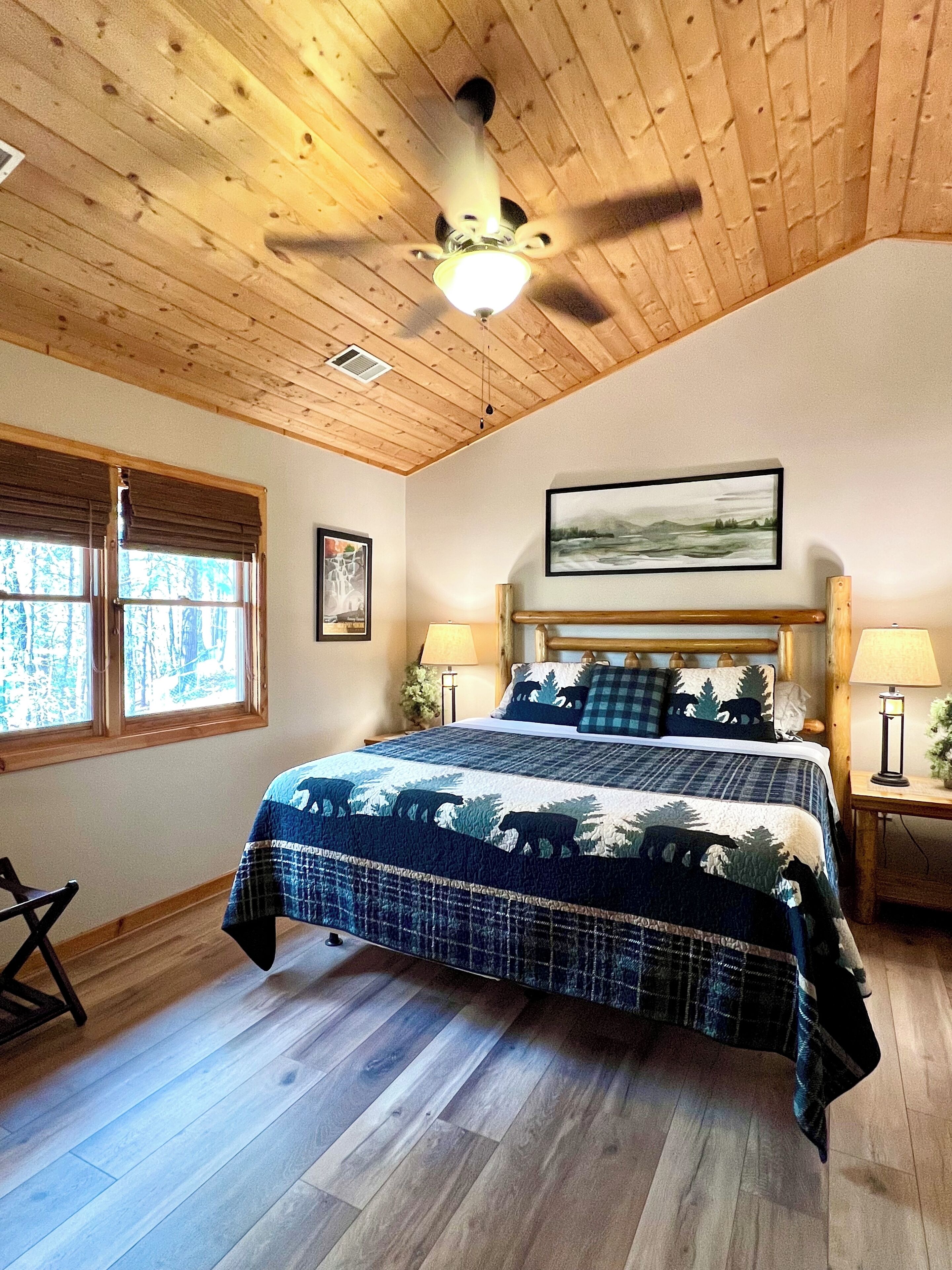 Una camera da letto con soffitto in legno, un letto con copriletto a tema foresta, due lampade ai piedi del letto e una finestra con persiane.