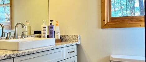 A bathroom with a white sink, vanity, and toilet. There are three bottles of lotion on the vanity, and a window with a view of trees.