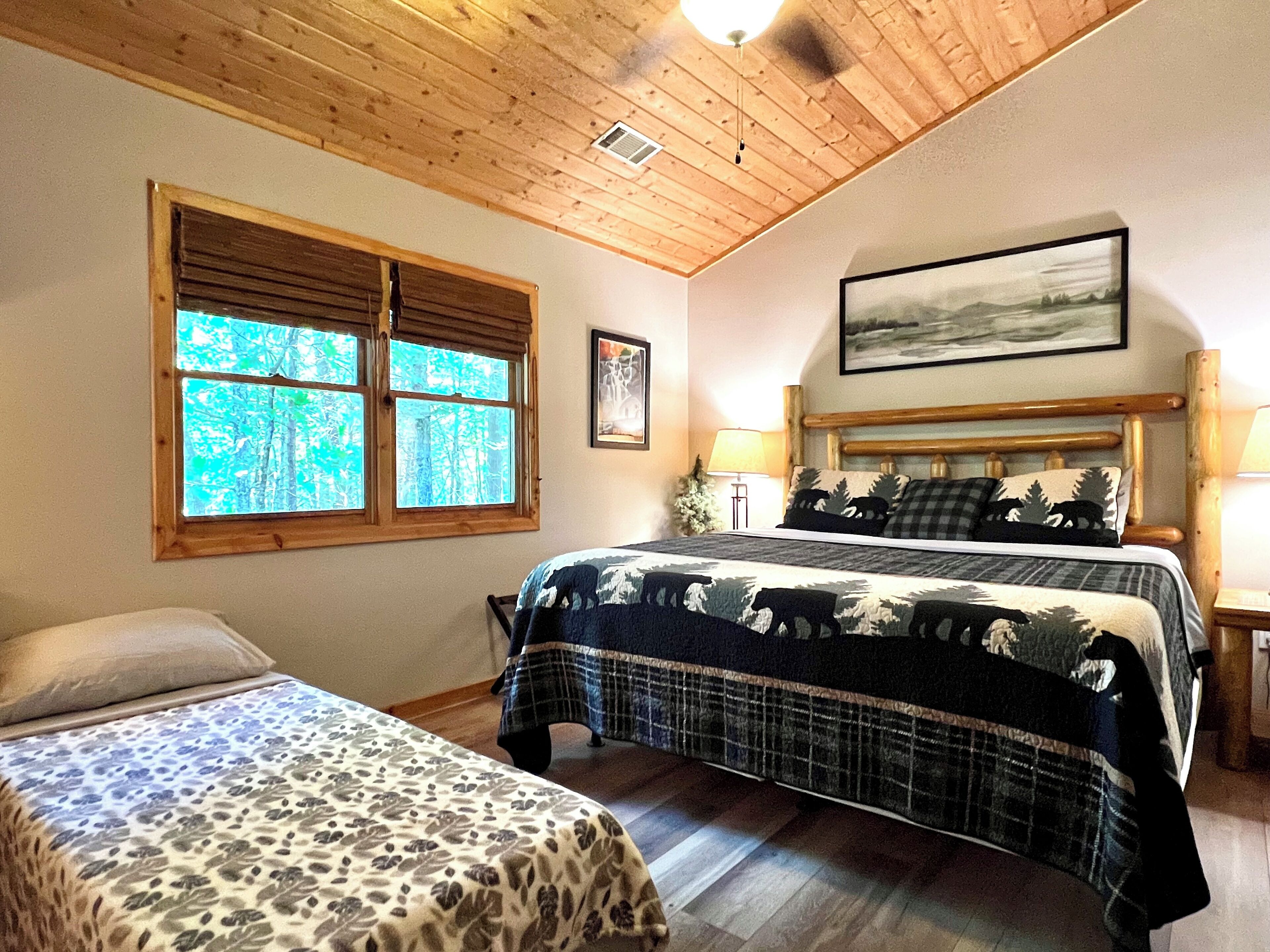 A bedroom with a large bed, a smaller bed, a window with a view of trees, a headboard with a painting, and a lamp on a bedside table.