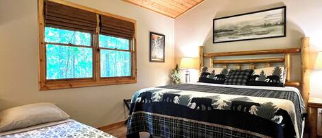 A bedroom with a large bed, a smaller bed, a window with a view of trees, a headboard with a painting, and a lamp on a bedside table.