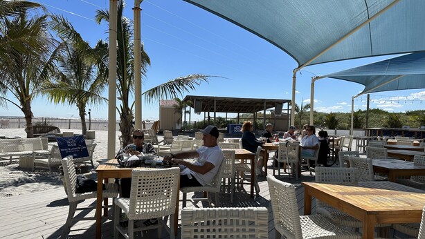 Un espacio al aire libre con sillas y mesas de mimbre, una zona con sombra y vista a la playa.