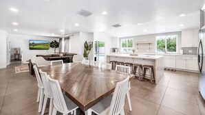 Uma cozinha ampla com armários brancos, uma ilha grande com banquetas e uma área de jantar com mesa de madeira e cadeiras brancas.