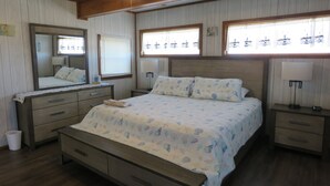 Un dormitorio con cabecera de madera, cama con colcha estampada, mesita de noche con lámpara, cómoda con cajones, espejo y ventilador de techo.