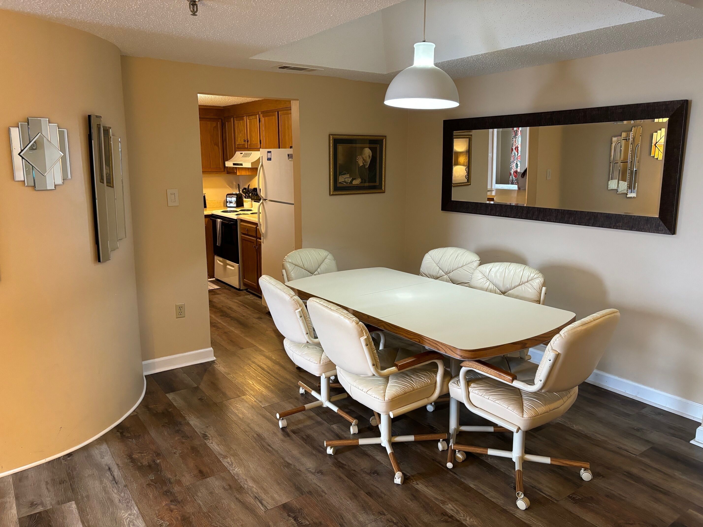 A dining area with a white table and leather chairs, a kitchen in the background, and a large mirror on the wall.