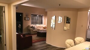 Una sala de estar con un sofá, un reposapiés y una mesa de comedor con sillas. Hay una ventana con persianas y una puerta que da a otra habitación.