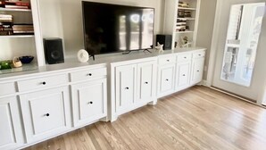 Une pièce spacieuse avec un grand téléviseur à écran plat fixé à un meuble blanc, un plancher en bois et une porte blanche donnant sur une autre pièce.