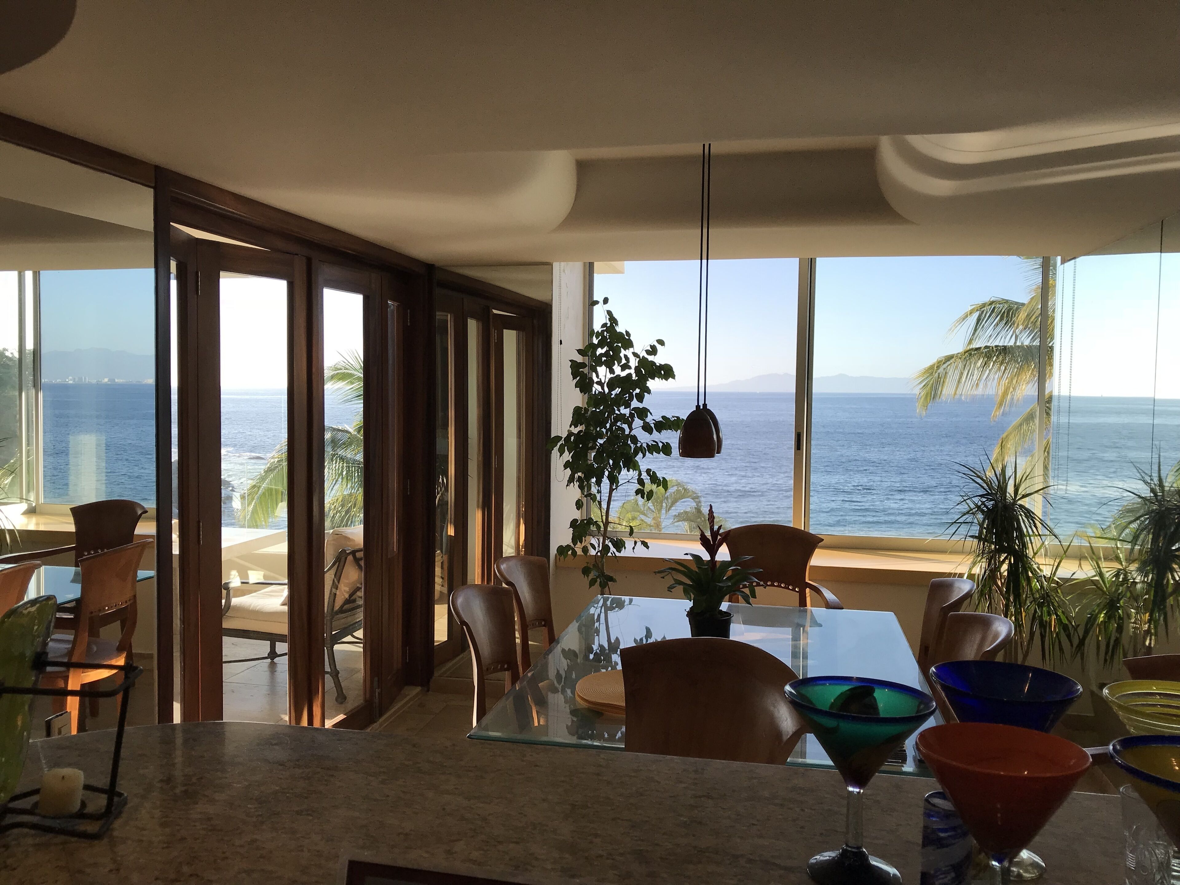Un comedor moderno con vista al océano, ventanas amplias y una variedad de cristalería colorida sobre la mesa.