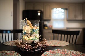Una mesa de comedor con un centro de mesa de vidrio transparente que contiene objetos decorativos e iluminación, rodeada de posavasos florales.