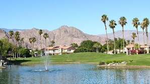 A golf course with a pond, palm trees, and houses in the background. - Absolutely Pristine 2-2 Lower Unit with Stunning Views! 2027 BNP Paribas Close!! (La Quinta)