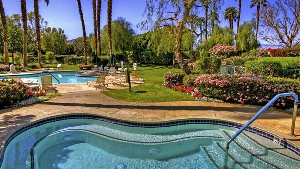A swimming pool with a tiled edge, surrounded by palm trees and a well-maintained garden with various plants and flowers. - Absolutely Pristine 2-2 Lower Unit with Stunning Views! 2027 BNP Paribas Close!! (La Quinta)