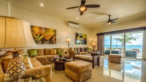 A spacious living room with a large sofa, ottoman, and coffee table. There are ceiling fans, a large window with a view of the ocean, and artwork on the walls. - Luxurious and elegant beachfront villa with a heated pool! (Akumal)