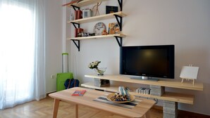 Ein Wohnzimmer mit Flachbildfernseher, Holzregalen, einem grünen Koffer und einem Tisch mit einer Schale Snacks.