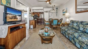 A living room with a television, a glass coffee table, a sofa, and a dining area in the background. - Maui Banyan P304 - Remodeled, Partial Ocean View, AC, Across from Kamaole Beach! (Kihei)
