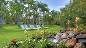 A lush garden with a small waterfall, palm trees, and two lounge chairs. - Maui Banyan P304 - Remodeled, Partial Ocean View, AC, Across from Kamaole Beach! (Kihei)