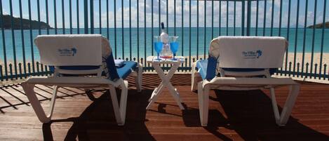 Deux fauteuils de jardin blancs avec des coussins bleus, placés sur une terrasse en bois, face à une mer turquoise, avec une clôture métallique en arrière-plan.