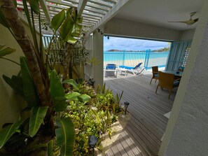 Una terraza de madera con mobiliario exterior, baranda blanca y plantas tropicales.
