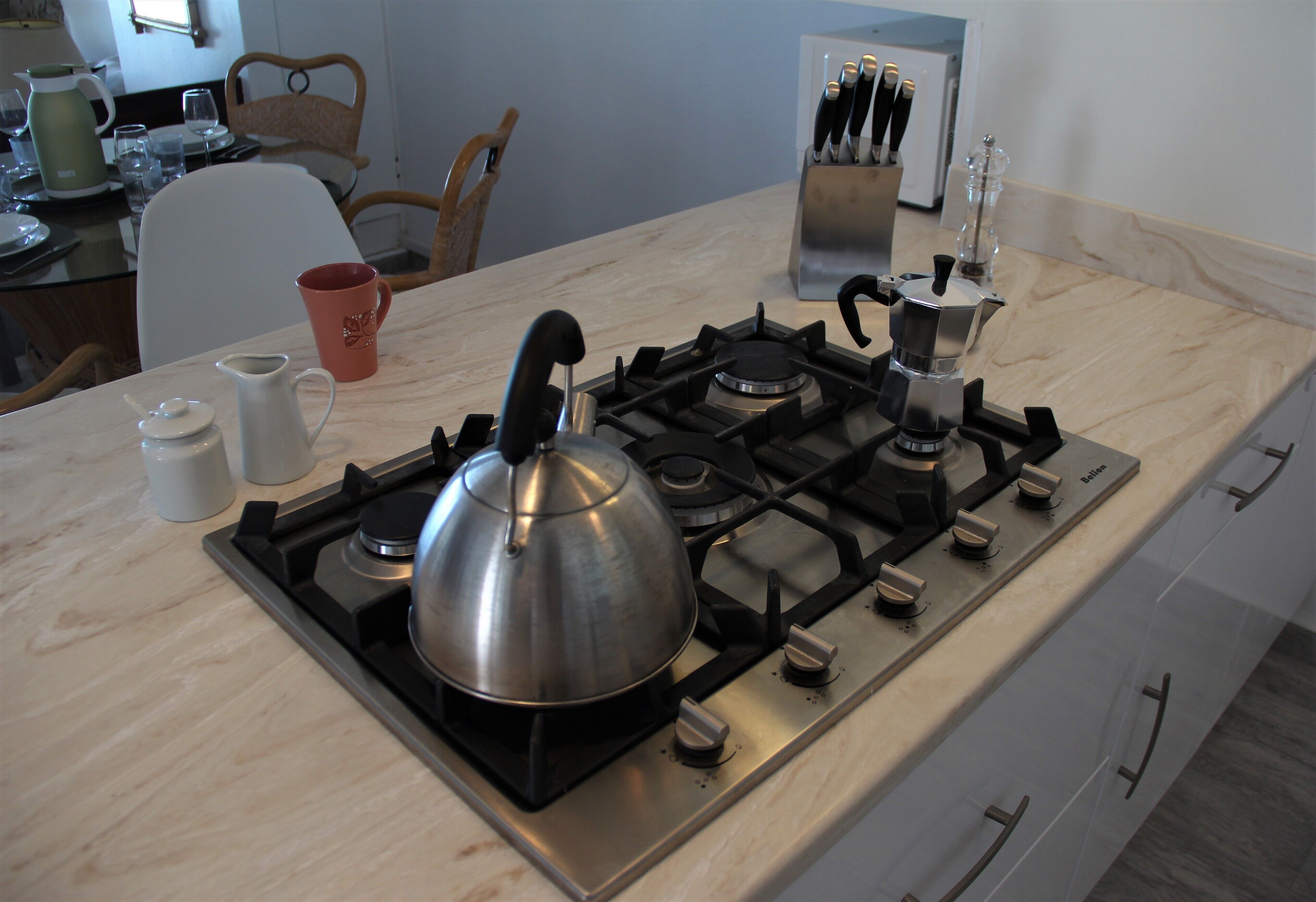 Cucina con piano cottura a gas, bollitore e macchina per il caffè. Sullo sfondo, sedie e un tavolo da pranzo.