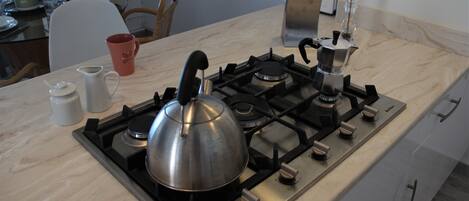 Cucina con piano cottura a gas, bollitore e macchina per il caffè. Sullo sfondo, sedie e un tavolo da pranzo.