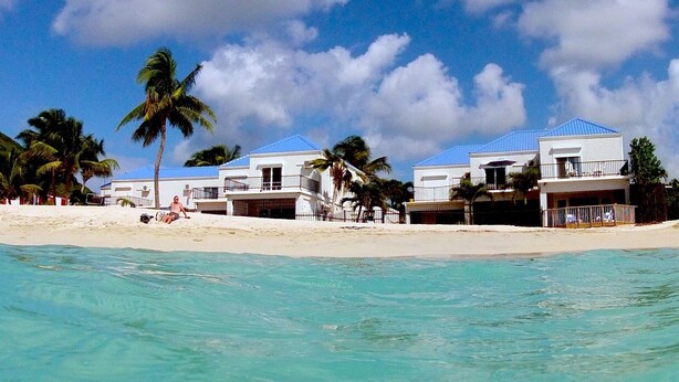 Una propiedad frente a la playa con varias edificaciones blancas, cada una con techo azul y balcones. Palmeras bordean la playa arenosa, y el océano se ve en primer plano.