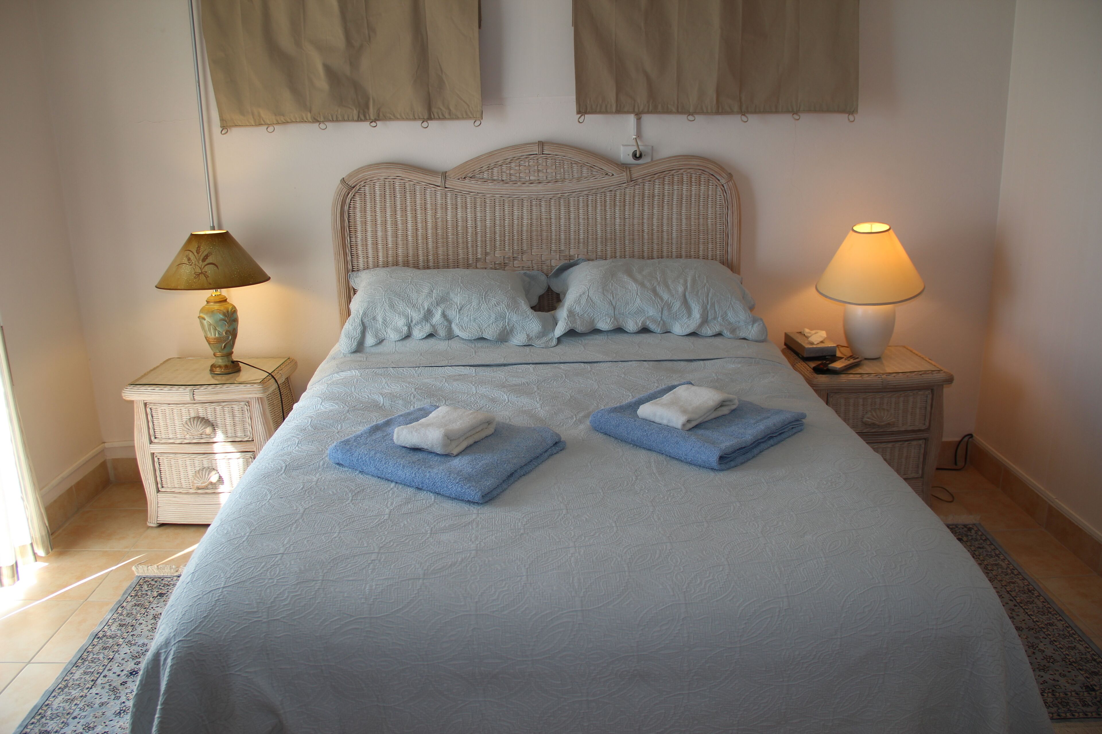 A neatly made bed with two pillows and two folded towels, a wicker headboard, two bedside tables with lamps, and a window with curtains.