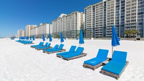 A beachside hotel with rows of blue sun loungers and umbrellas, large buildings in the background, and a clear blue sky.
