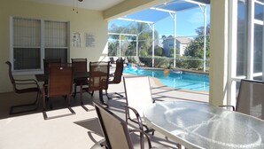 Un patio iluminado por el sol con una mesa y sillas de vidrio, un ventilador de techo y vistas a una piscina y casas.