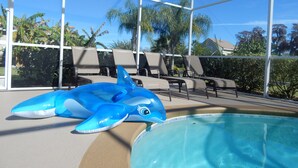 Un área de piscina interior con un juguete inflable con forma de delfín, sillas de descanso y una vista despejada de palmeras y casas al fondo.