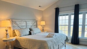 Ein ordentlich bezogenes Bett mit gesteppter Decke, zwei Nachttischlampen und Blick ins Freie durch ein Fenster mit Vorhängen.