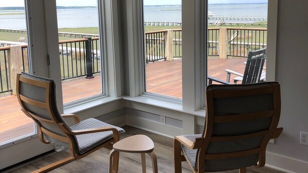 Ein Zimmer mit zwei Holzstühlen und einem kleinen Tisch, große Fenster mit Blick auf ein Gewässer und eine entfernte Küste.