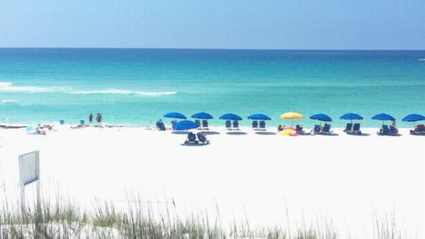 Une plage de sable blanc bordée d’une eau d’un bleu cristallin. Plusieurs parasols et des chaises de plage sont disposés le long du rivage.