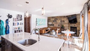 A modern kitchen with a large island, a dining area with a round table, a stone fireplace, and a flat-screen TV mounted on the wall. - Minutes to festivals - Pool, Jacuzzi, Fitness room (Snowmass Village)