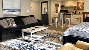 A compact living space with a sofa, a coffee table, a patterned rug, a bed, a kitchen area with stools, and a large painting on the wall. - In town, newly remodeled studio with private balcony  (Shelburne Falls)