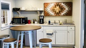 A compact kitchen with white cabinets, a microwave, a coffee maker, and a decorative wooden wall piece. There are two stools and a small table. - In town, newly remodeled studio with private balcony  (Shelburne Falls)