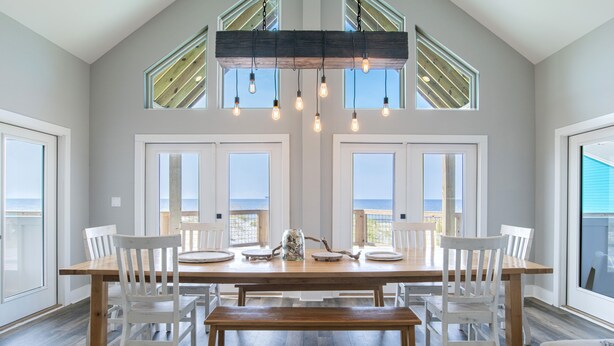 A dining area with a large wooden table, white chairs, and a bench. There are multiple windows providing natural light and views of the outdoors. - Amazing Views! Beachfront Home Sleeps 16-Private Pool & Private Beach! (Gulf Shores)