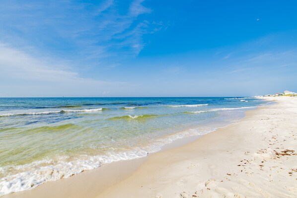 A beach with clear blue water, sandy shore, and a few buildings in the distance.