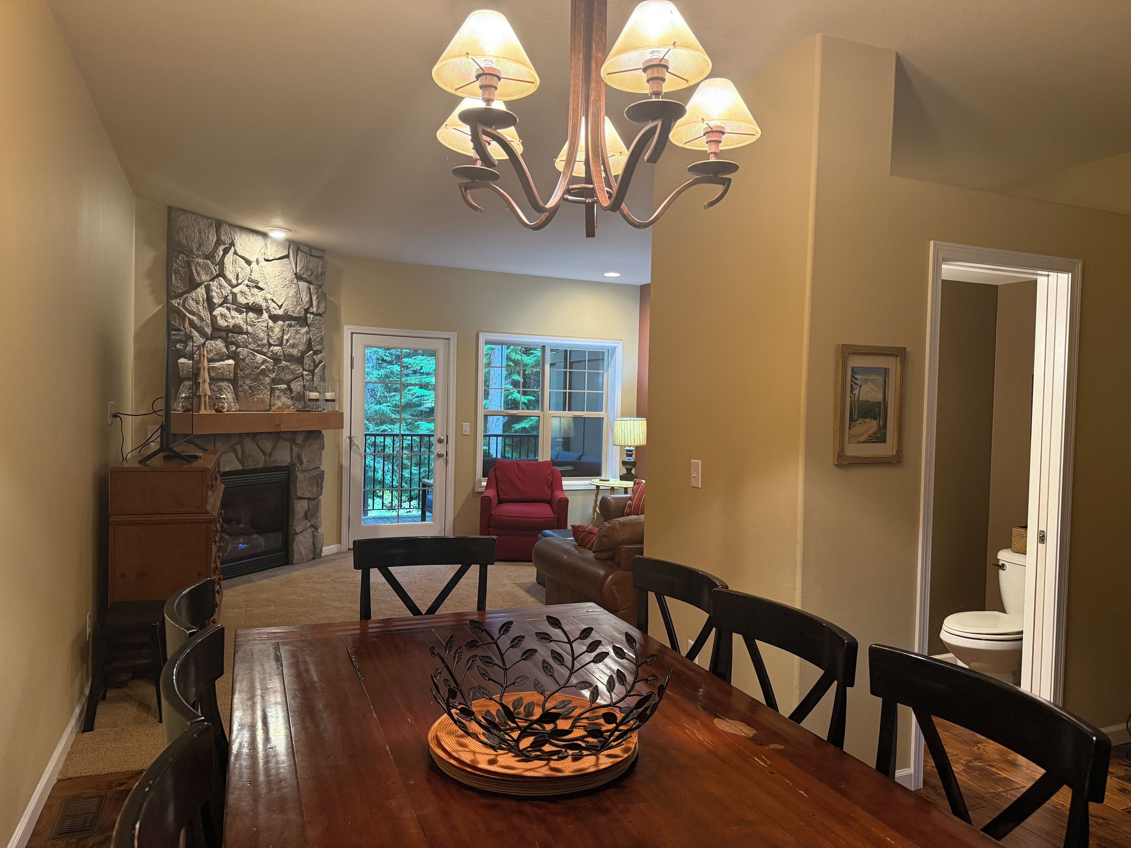 A dining area with a wooden table, chairs, a chandelier, a fireplace, and a view of a forest through a glass door.