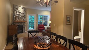 A dining area with a wooden table, chairs, a chandelier, a fireplace, and a view of a forest through a glass door. - Ideal location in Collins Lake, close to pool with views of Forest & the Slopes (Government Camp)