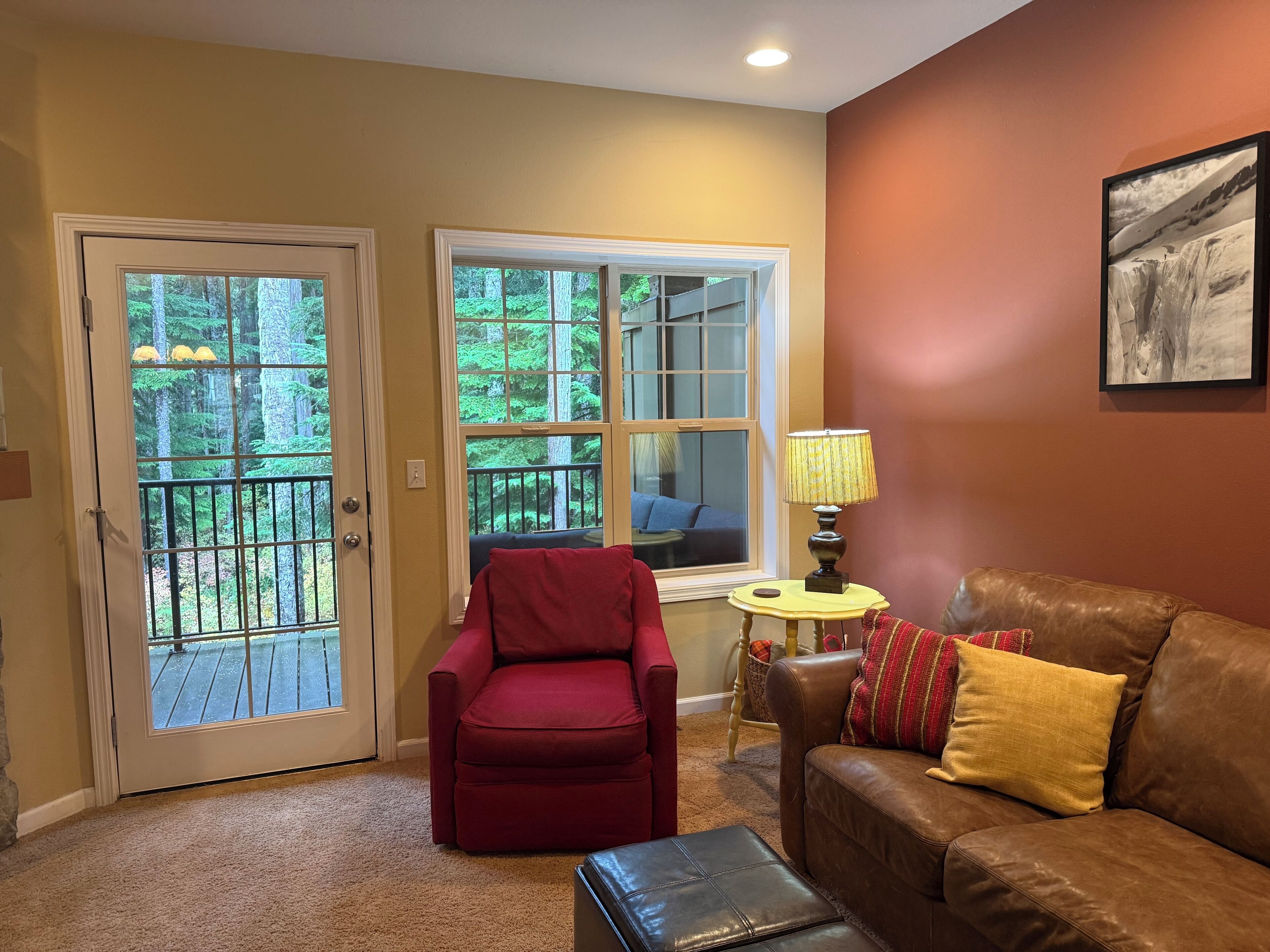 A living room with a red armchair, a brown leather couch, and a small round table with a lamp. There are two windows with a view of a deck and trees.