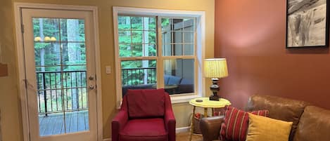 A living room with a red armchair, a brown leather couch, and a small round table with a lamp. There are two windows with a view of a deck and trees.