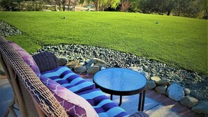 A wicker lounge chair with cushions, a small round table, and a view of a grassy area with trees and a distant mountain range. - MYSTICAL HILLTOP RANCH (Santa Ynez)