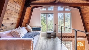 Ein gemütliches Schlafzimmer im Dachgeschoss mit Holzbalkendecke, einem Bett mit Kissen, einem Nachttisch und einem großen Fenster mit Blick auf Bäume.