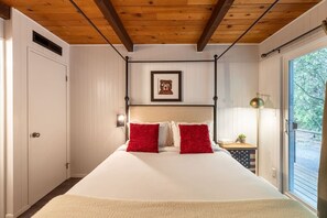 A bedroom with a large bed, white walls, wooden ceiling beams, a framed picture above the bed, and a window with a view of trees. - Cozy A-Frame w/Spa+14 acres+EV+Trails+Lake Access (Lakeside)