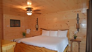 A wooden cabin bedroom with a bed, a ceiling fan, a wall-mounted lamp, a small table with a plant, and a framed picture on the wall. - Luxury 4 King Suites, 4 Bath Log Cabin Close to Town & Trolley (Gatlinburg)