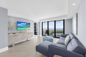 A modern living room with a large flat-screen TV, grey sofa, and a view of the sea through large windows.
