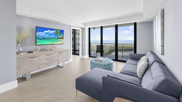 Ein modernes Wohnzimmer mit großem Flachbildfernseher, grauem Sofa und Meerblick durch hohe Fenster.