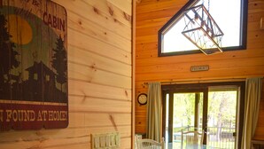 A cabin interior with a wooden wall, a dining table with chairs, a hanging light fixture, and a large window.
