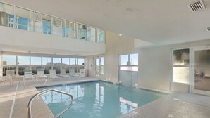 Une piscine intérieure dotée de rampes en verre transparent, un espace aménagé au bord de la piscine avec des chaises longues, et une vue sur les gratte-ciel de la ville à travers de grandes fenêtres.