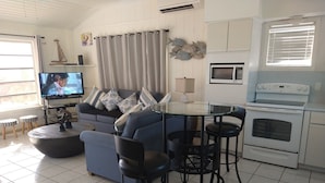 A living room with a blue sofa, a glass coffee table, black chairs, a TV, a kitchen area with a microwave and stove, and a window with blinds.