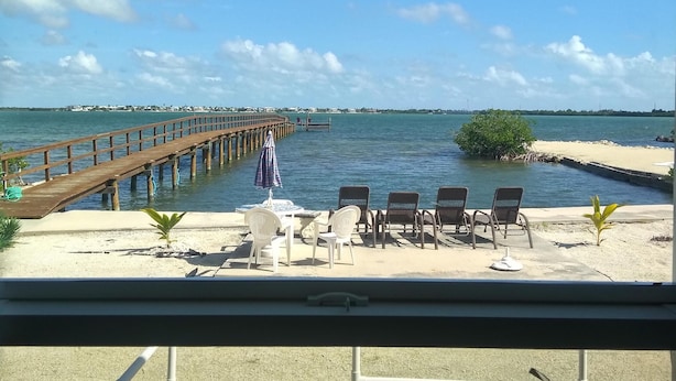 A beachside view with a wooden pier extending into the water, a sandy area with lounge chairs and umbrellas, and a clear blue sky.