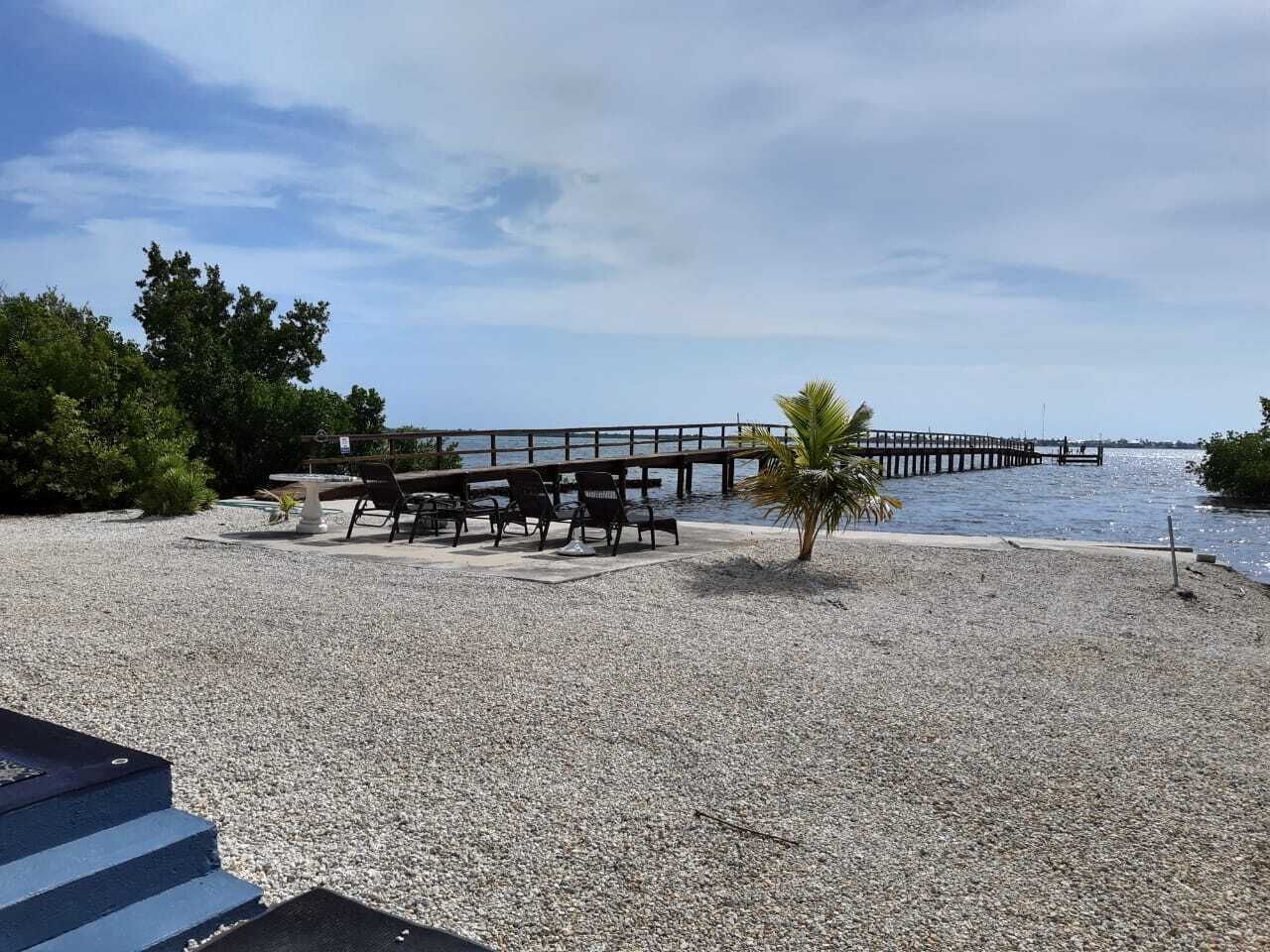A pebbled beach area with a wooden pier extending into the water, surrounded by greenery and a clear sky.